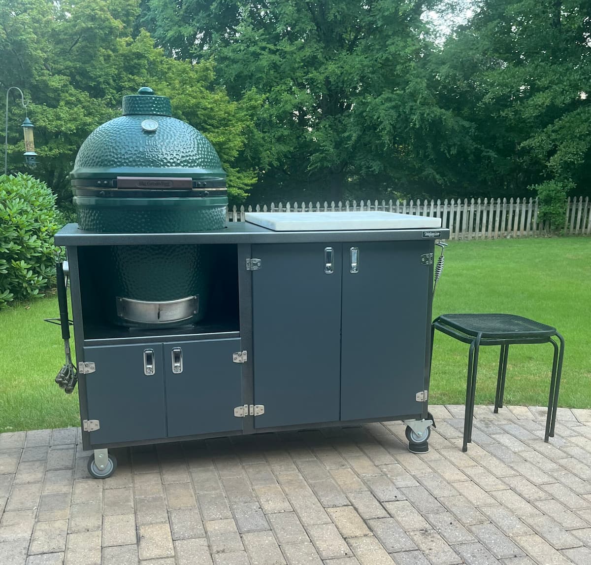 The image shows a green ceramic Big Green Egg grill installed within a dark gray outdoor kitchen cabinet on a paved patio. The cabinet features storage doors, metal handles, and caster wheels for mobility, with grilling tools hanging on the side. In the background, a neatly maintained lawn, white picket fence, and tall leafy trees create a peaceful backyard setting.